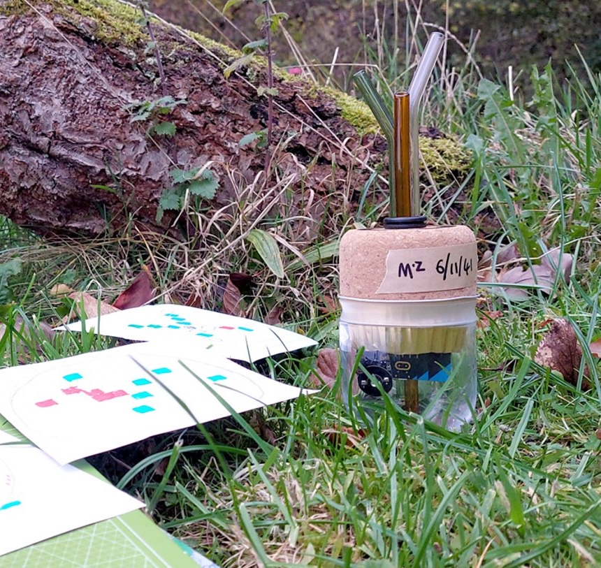 A mysterious cork and plastic onject intended to represent a soil health sensor, surrounded by wood, grass, and pieces of paper