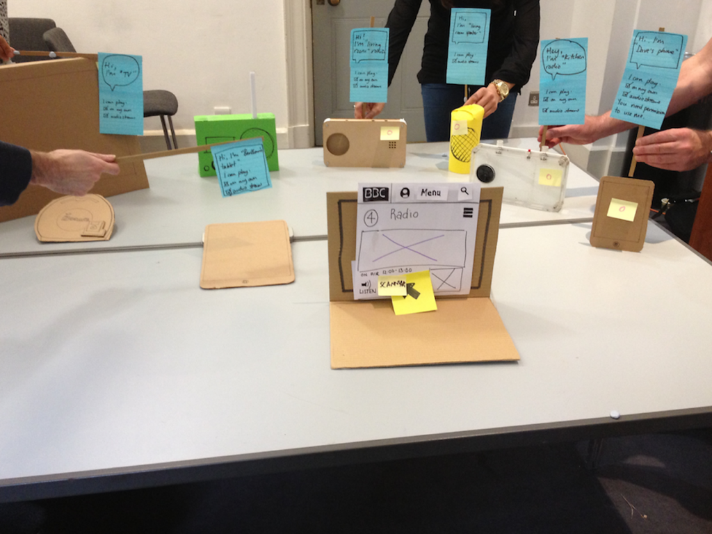 Various carboard versions of devices with little postit note signs above them held by humans explainig what they are doing